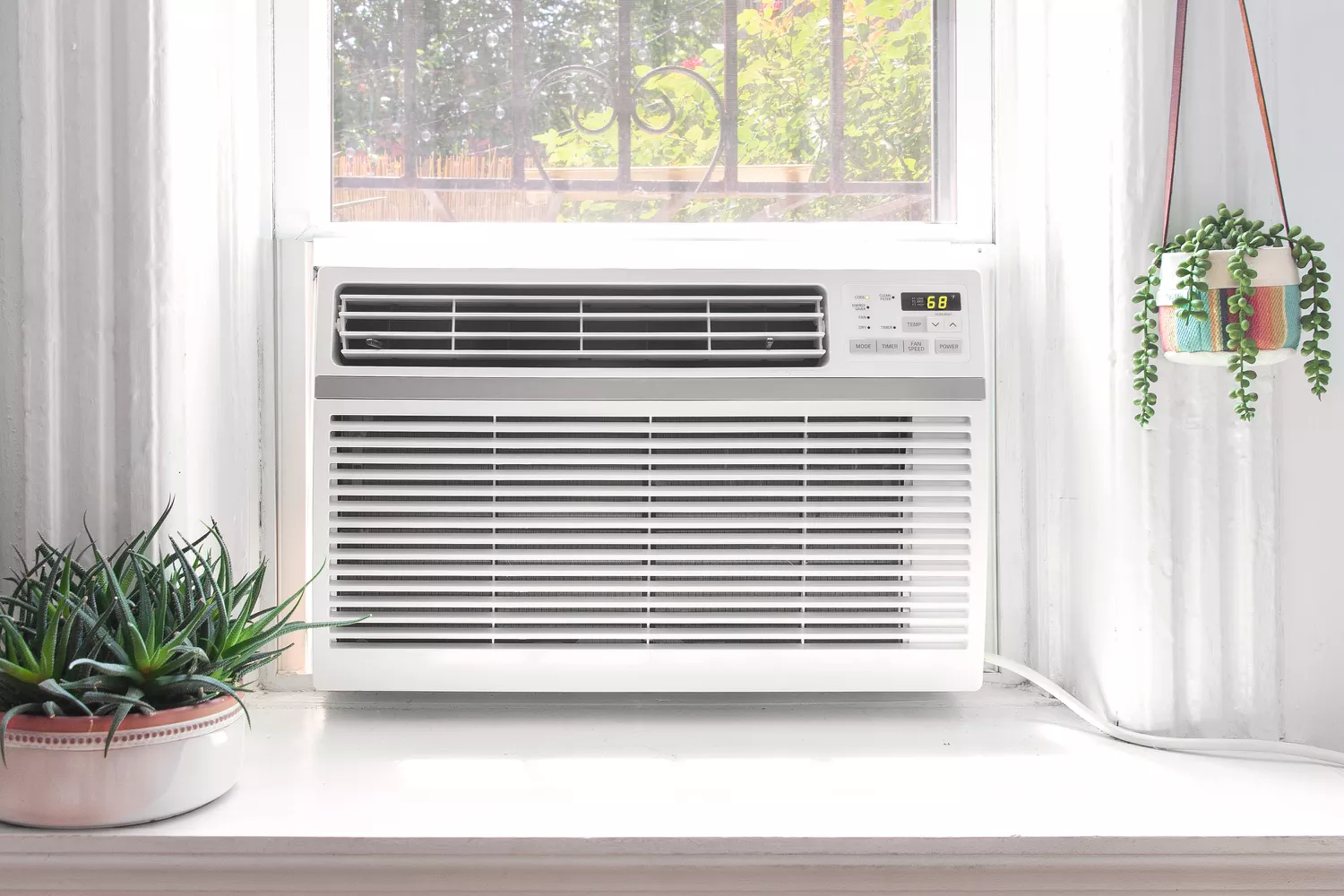 Window-mounted air conditioner surrounded by indoor plants, placed under a window with sunlight